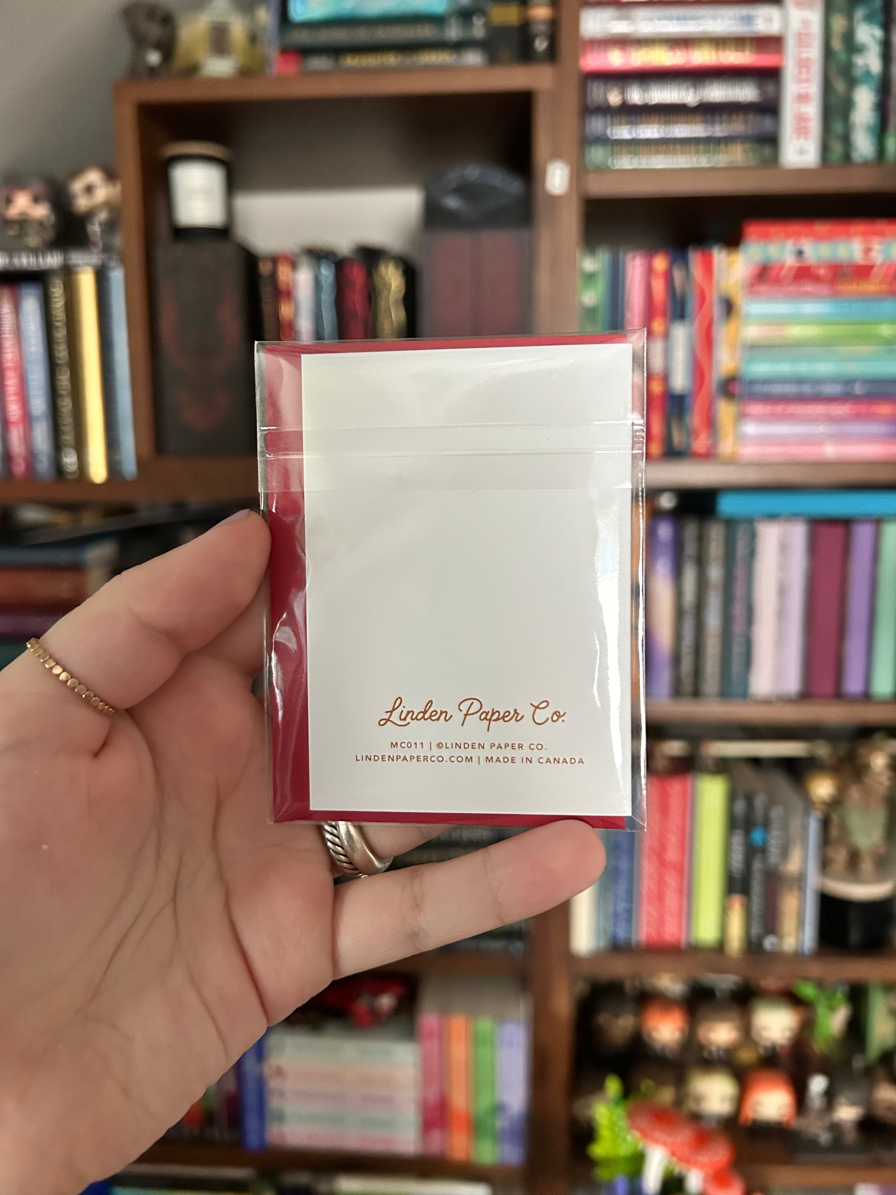 A hand holding up the back of a Christmas card, printed with the brand name 'Linden Paper Co.' in gold, against a blurred background of bookshelves.