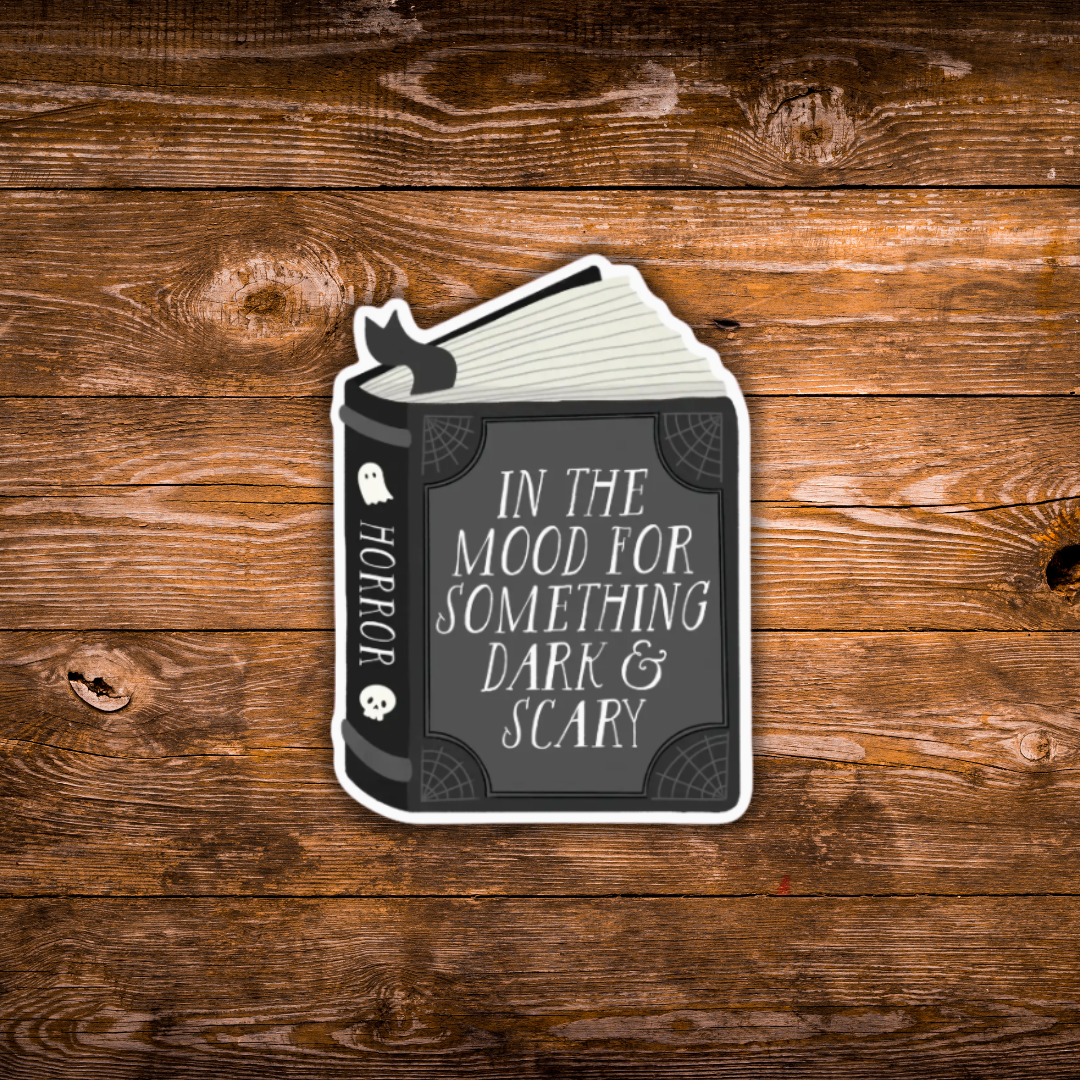 A die-cut sticker shaped like an open horror book with the text 'IN THE MOOD FOR SOMETHING DARK & SCARY' on the front cover and 'HORROR' on the spine, shown on a rustic brown wood plank background.