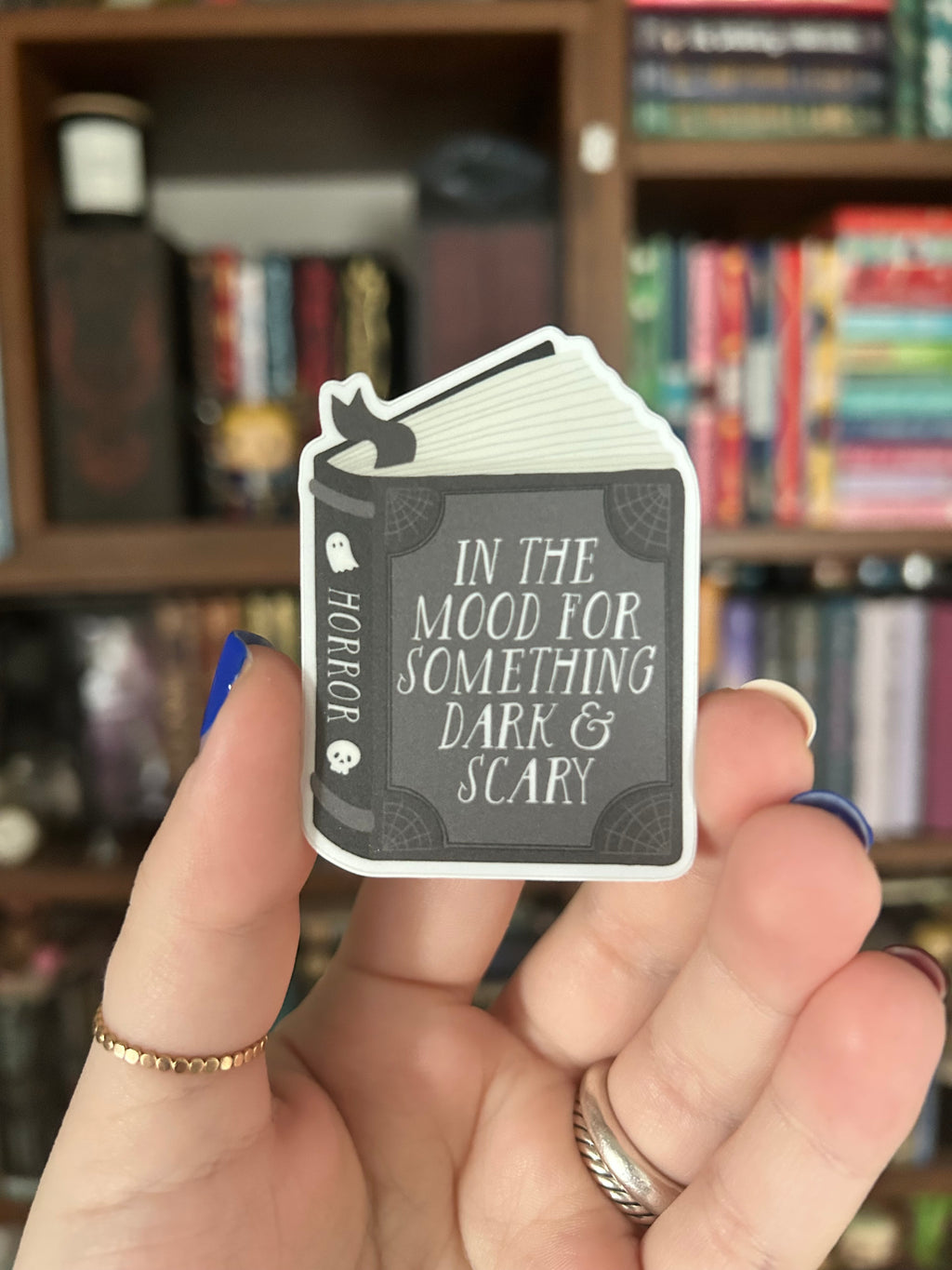 A hand holding a die-cut sticker shaped like a book cover with the text 'IN THE MOOD FOR SOMETHING DARK & SCARY' and 'HORROR' on the cover, with a blurred bookshelf in the background.