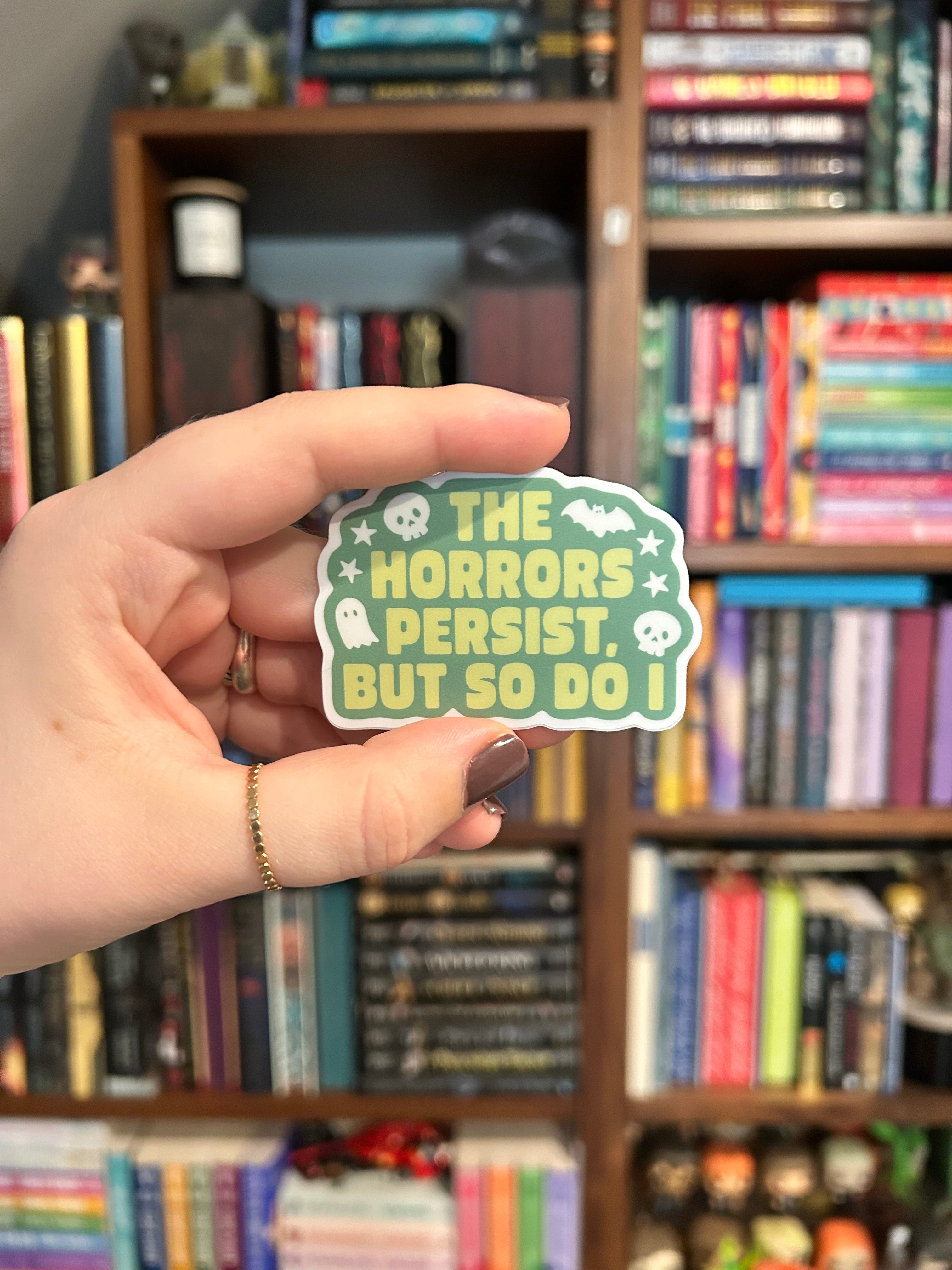 A hand holds a die-cut sticker featuring the text 'THE HORRORS PERSEST. BUT SO DO I' in yellow on a green background with white skull and bat illustrations.
