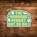 A die-cut sticker with the phrase 'THE HORRORS PERSIST, BUT SO DO I' in pale yellow text on a mint green background, decorated with white skulls, a bat, stars, and a ghost. The sticker is displayed flat on a rustic brown wooden surface.