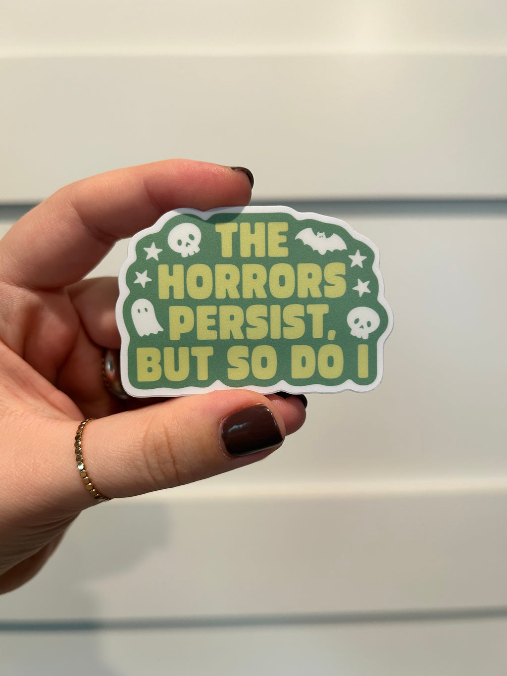 A hand holds a die-cut sticker featuring the text 'THE HORRORS PERSEST. BUT SO DO I' in yellow on a green background with white skull and bat illustrations.