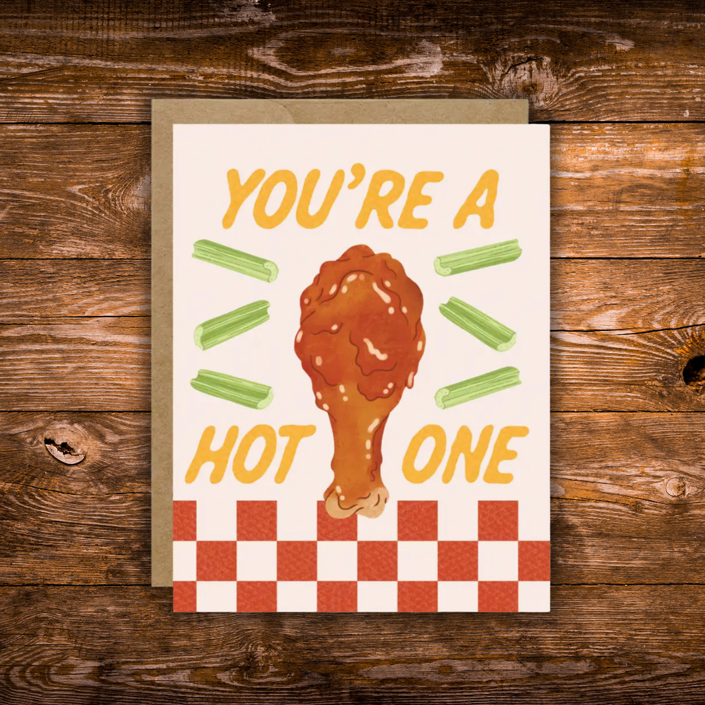 A square greeting card with a brown envelope, featuring a chicken leg illustration and the text 'YOU'RE A HOT ONE' with celery sticks, placed on a rustic wooden surface.