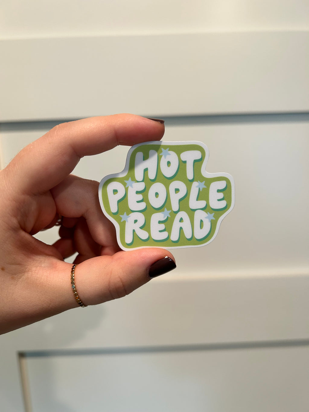A die-cut sticker with the phrase 'HOT PEOPLE READ' in white bubble letters on a light green background with small blue star accents