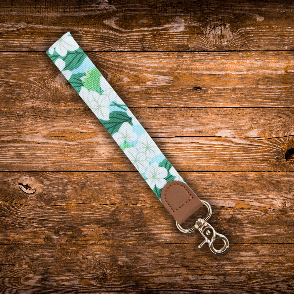 A fabric wristlet strap with a green and white Hydrangeas pattern, a brown faux-leather end tab, and a gold metal swivel clasp, laid diagonally on a rustic brown wooden background.