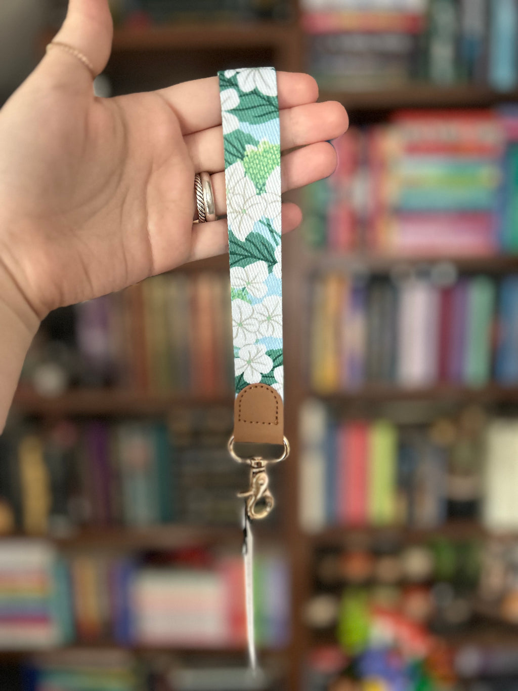 A hand holding a floral-patterned fabric wrist strap keychain with a light brown leather tab and gold-tone metal clasp, set against a blurred background of bookshelves.