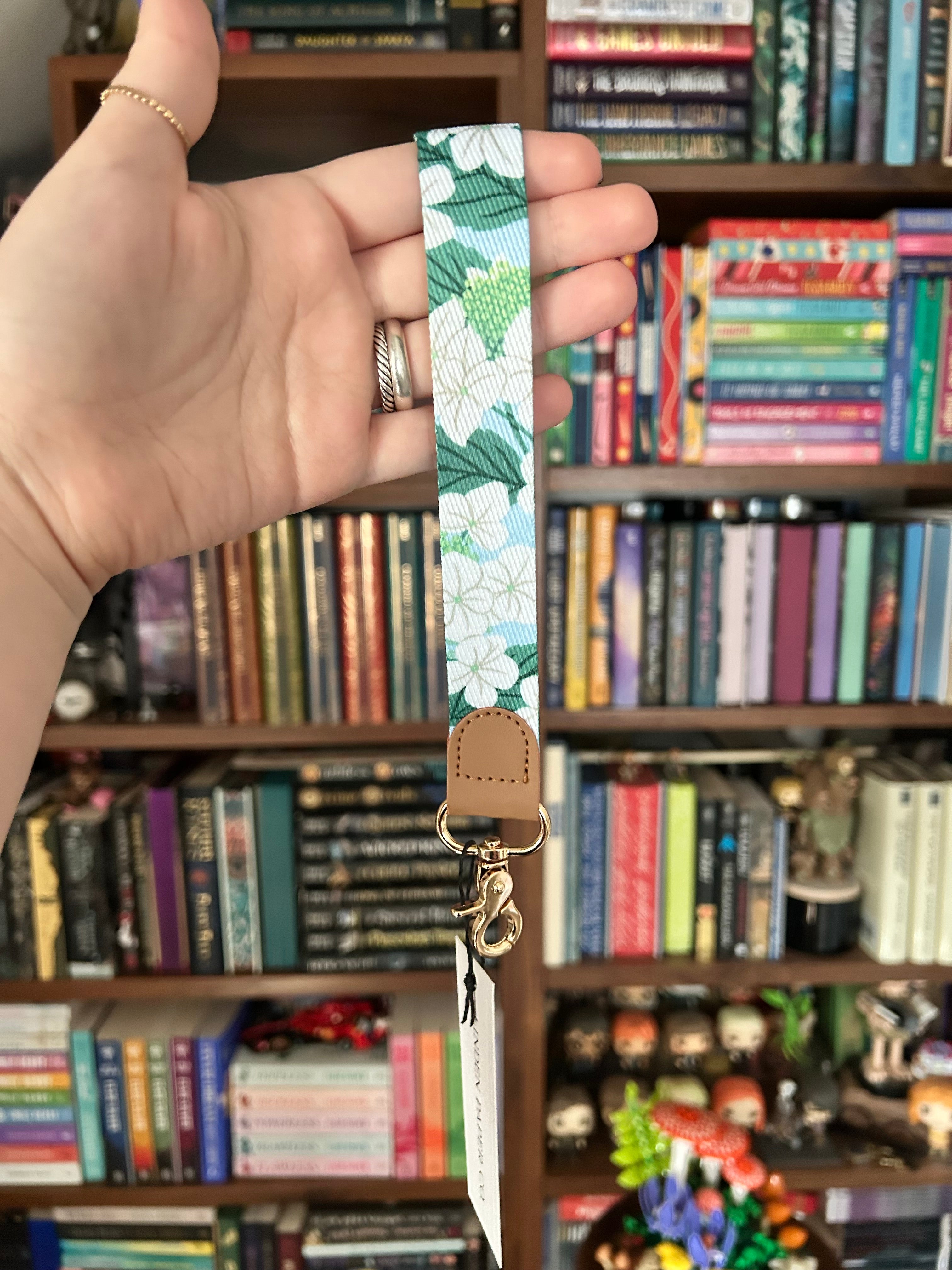 A hand holding a floral-patterned wrist strap with a green and white print, tan leather tab, and gold-toned metal clasp, set against a blurred background of bookshelves.
