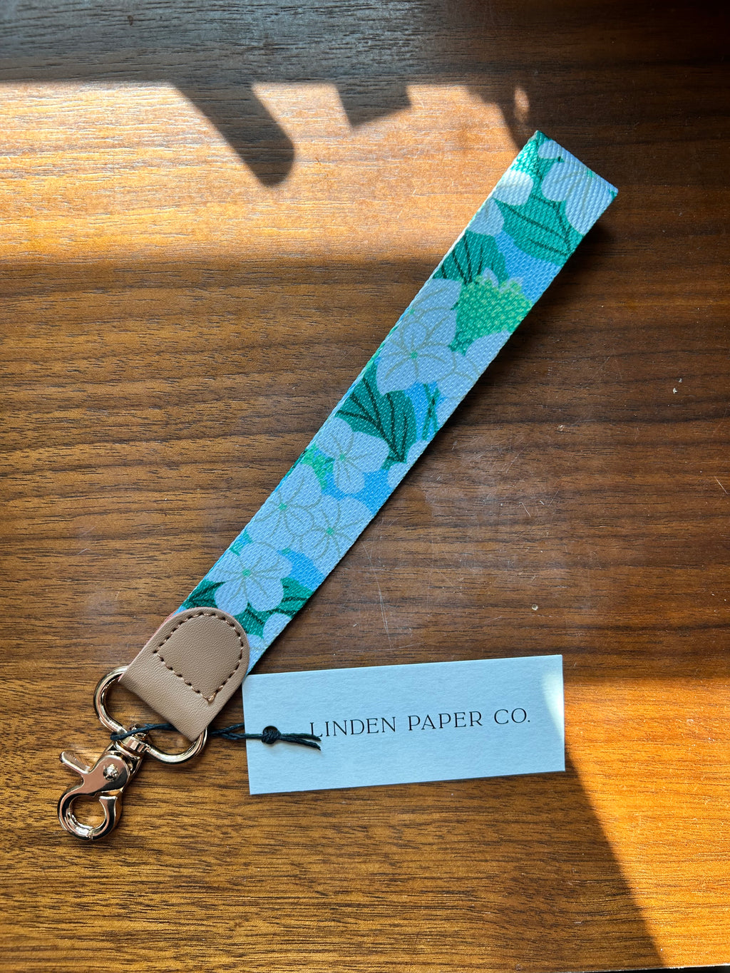 A fabric wrist strap keychain with a light blue and green floral pattern, a tan leather tab, and a silver metal clasp, shown on a wooden table next to a paper tag reading 'LINDEN PAPER CO'.