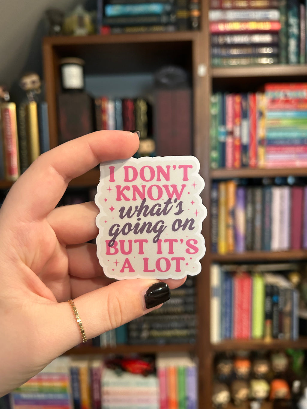 A hand with black nail polish holds a scalloped white sticker featuring the quote 'I DON'T KNOW what's going on BUT IT'S A LOT' in pink and dark gray lettering, with small pink stars. The background is a blurred bookshelf.