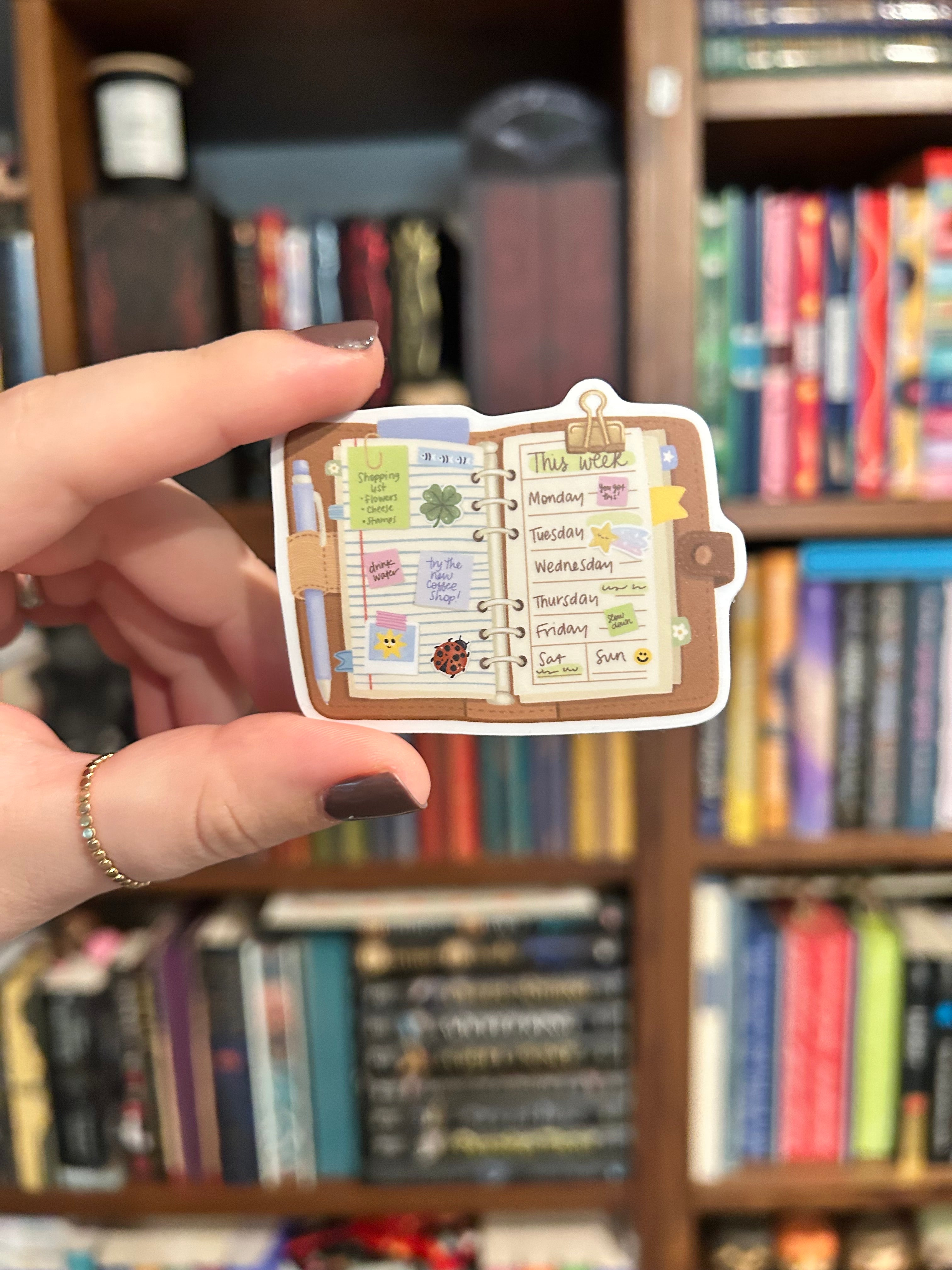 A hand with dark brown nail polish holds a small die-cut sticker shaped like an open planner or recipe book with colorful labels and illustrations, set against a blurred background of bookshelves.