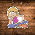 A cartoon die-cut sticker of a blonde girl with glasses sitting cross-legged, wearing a pink top and blue pants, placed on a brown wooden plank background.