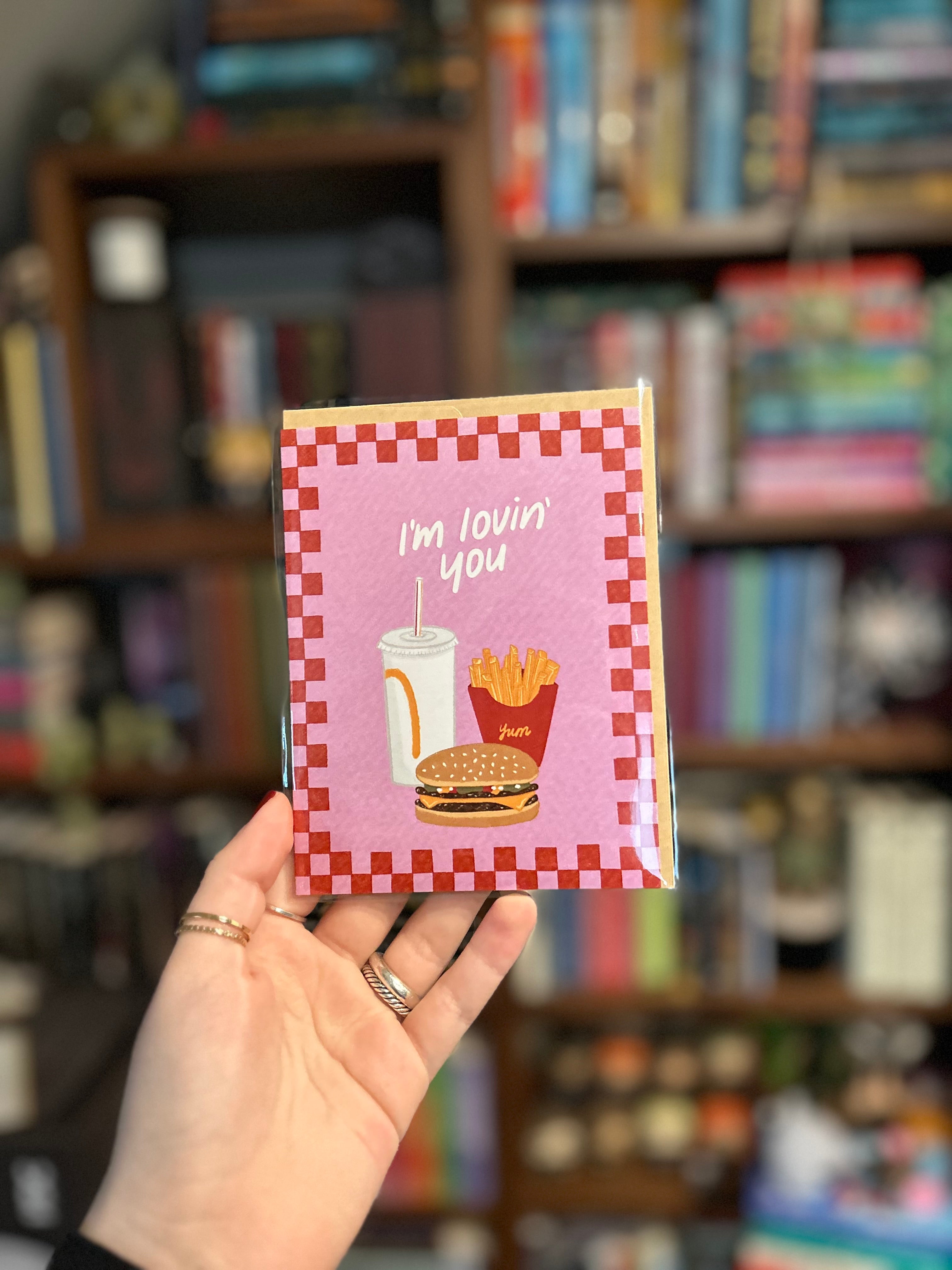 Cute romantic greeting card featuring an illustrated burger, fries, and soda with the message "I'm Lovin' You" on a retro pink background with a checkerboard border.