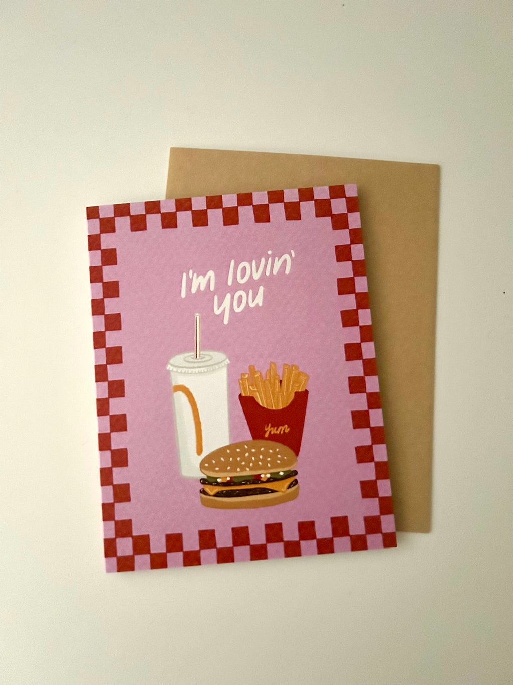 Cute romantic greeting card featuring an illustrated burger, fries, and soda with the message "I'm Lovin' You" on a retro pink background with a checkerboard border.