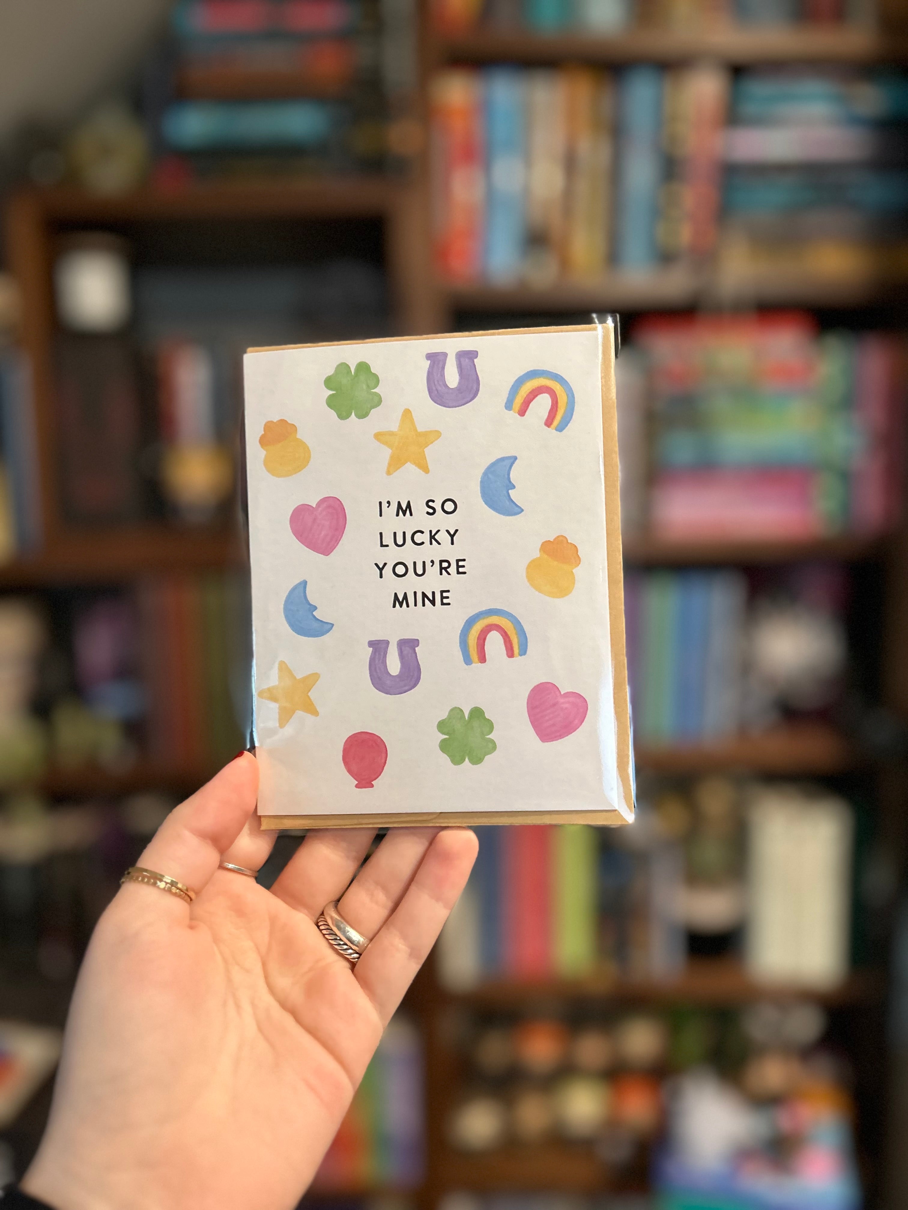 Cute romantic greeting card featuring colorful lucky charms including four-leaf clovers, horseshoes, rainbows, stars, hearts, and moons surrounding the message "I'm So Lucky You're Mine."