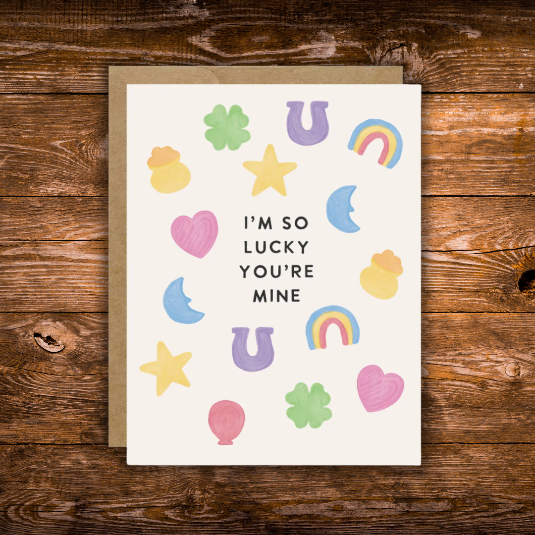 Cute romantic greeting card featuring colorful lucky charms including four-leaf clovers, horseshoes, rainbows, stars, hearts, and moons surrounding the message "I'm So Lucky You're Mine."