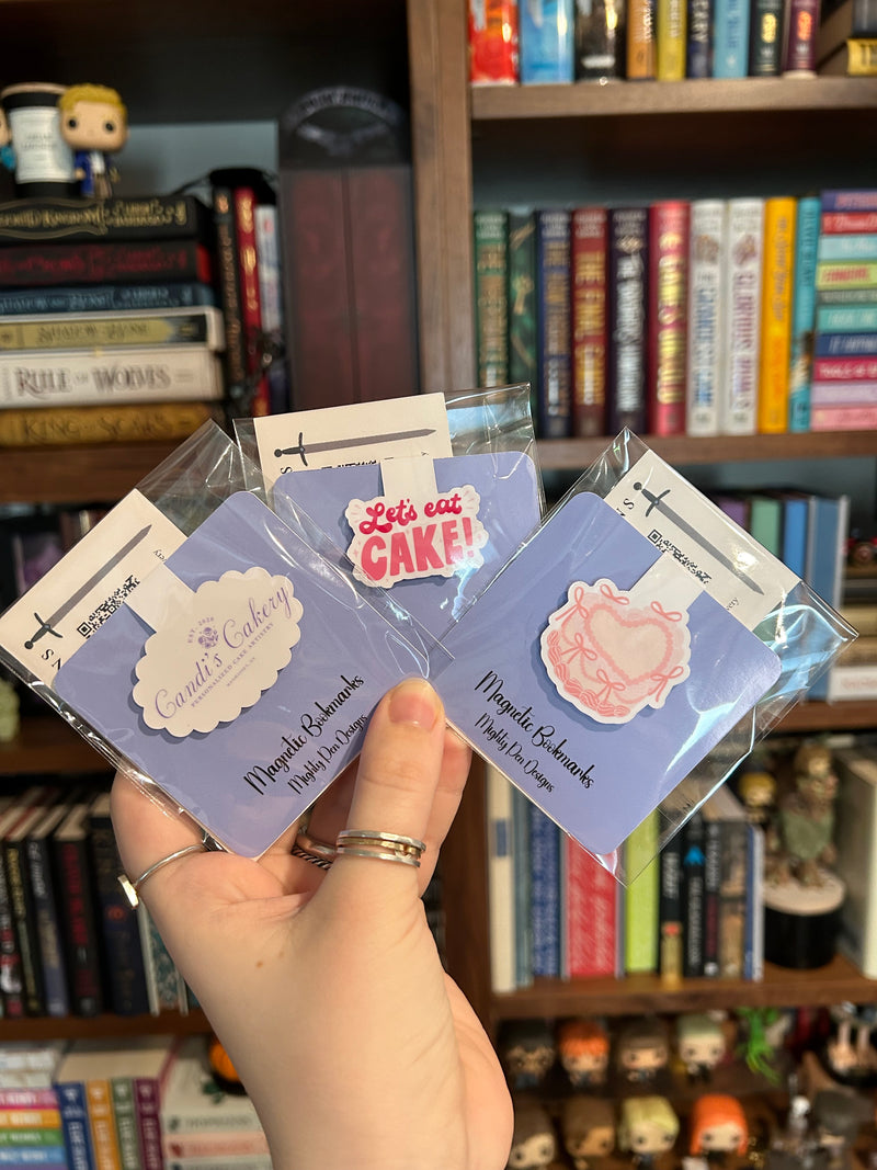 A hand holding three small magnet bookmarks in front of a bookshelf. The bookmarks are on lavender cards with decorative labels, including one that reads 'Let's eat CAKE!' and others with pastel illustrations.