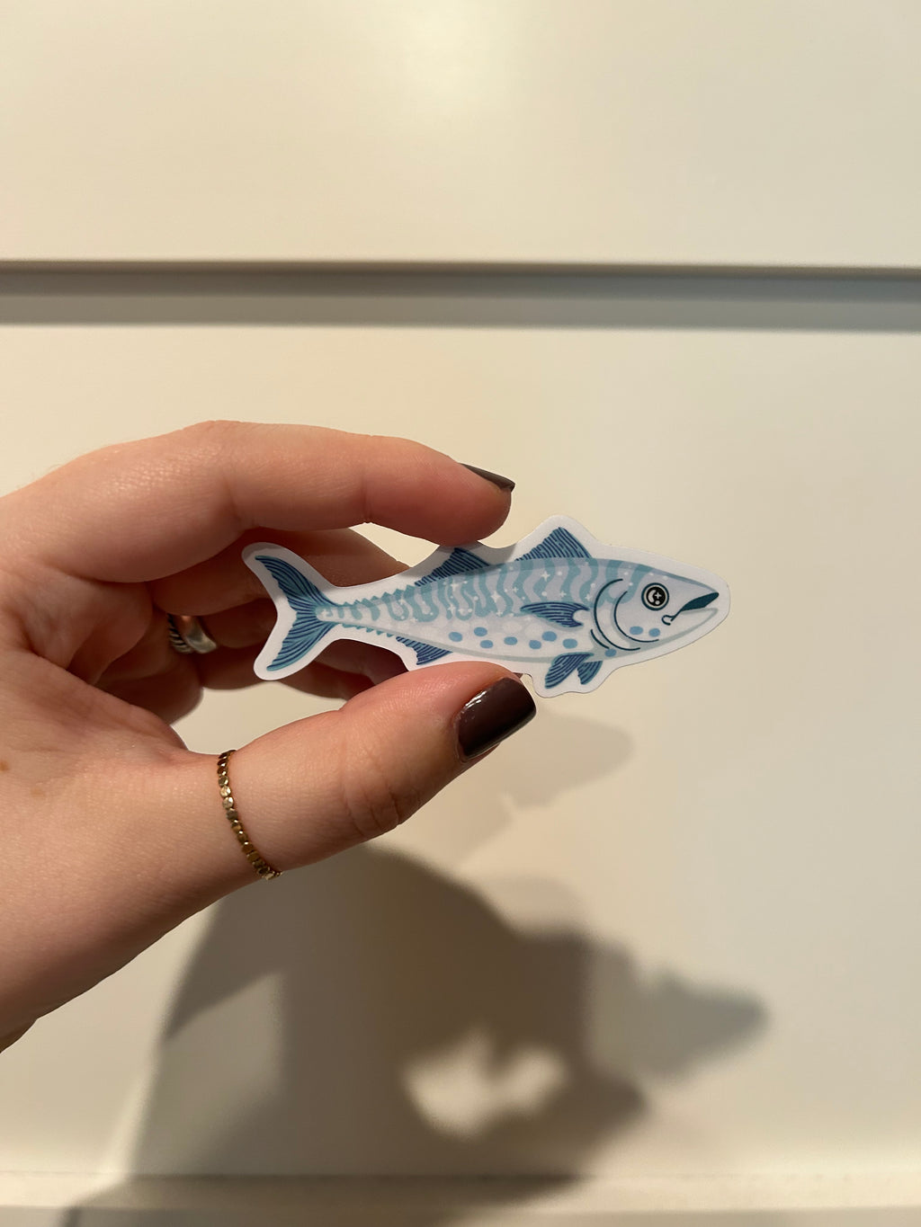 A hand with dark nail polish holds a die-cut sticker shaped like a blue and white spotted fish against a warm beige wall with a strong shadow.