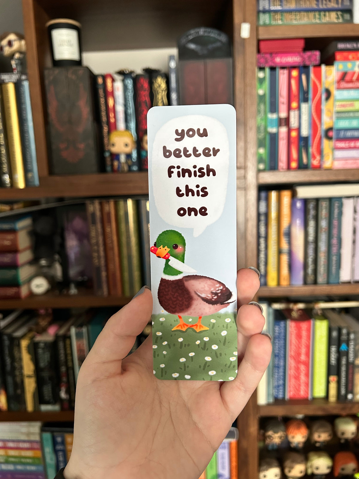 Double-sided mallard duck bookmark featuring a green-headed duck holding a tiny knife beneath the words “You better finish this one,” standing in a daisy meadow, with a white heart pattern on a light blue reverse.