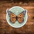 A die-cut circular sticker featuring an orange and black monarch butterfly on a pale green background with the words 'YOU ARE WHAT YOU MANIFEST' and small white stars and a crescent moon, shown on a rustic brown wood surface.