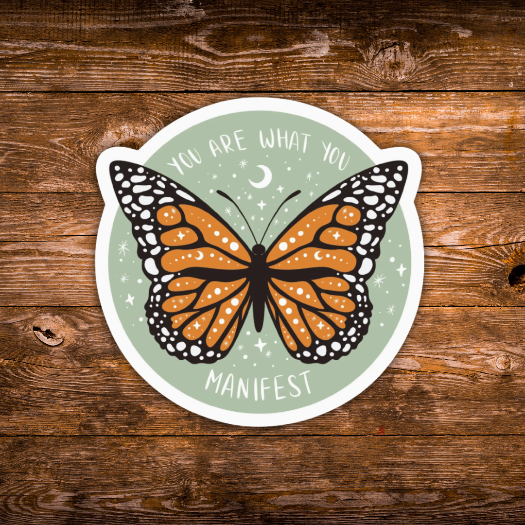 A die-cut circular sticker featuring an orange and black monarch butterfly on a pale green background with the words 'YOU ARE WHAT YOU MANIFEST' and small white stars and a crescent moon, shown on a rustic brown wood surface.