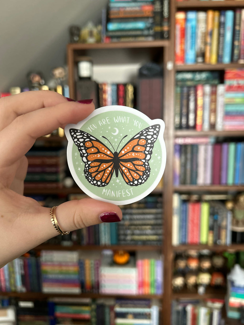 A hand with dark red nail polish holds a round sticker featuring an orange and black monarch butterfly on a pale green background with the text 'You Are What You Manifest' in a blurred bookshelf background.