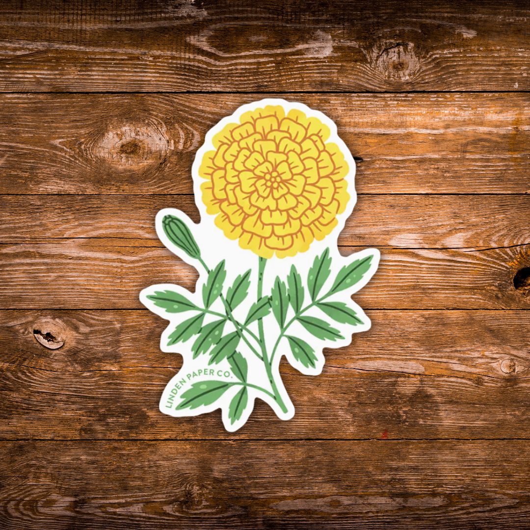 A die-cut sticker of a yellow marigold flower with green leaves on a white background, placed on a rustic brown wooden surface. 