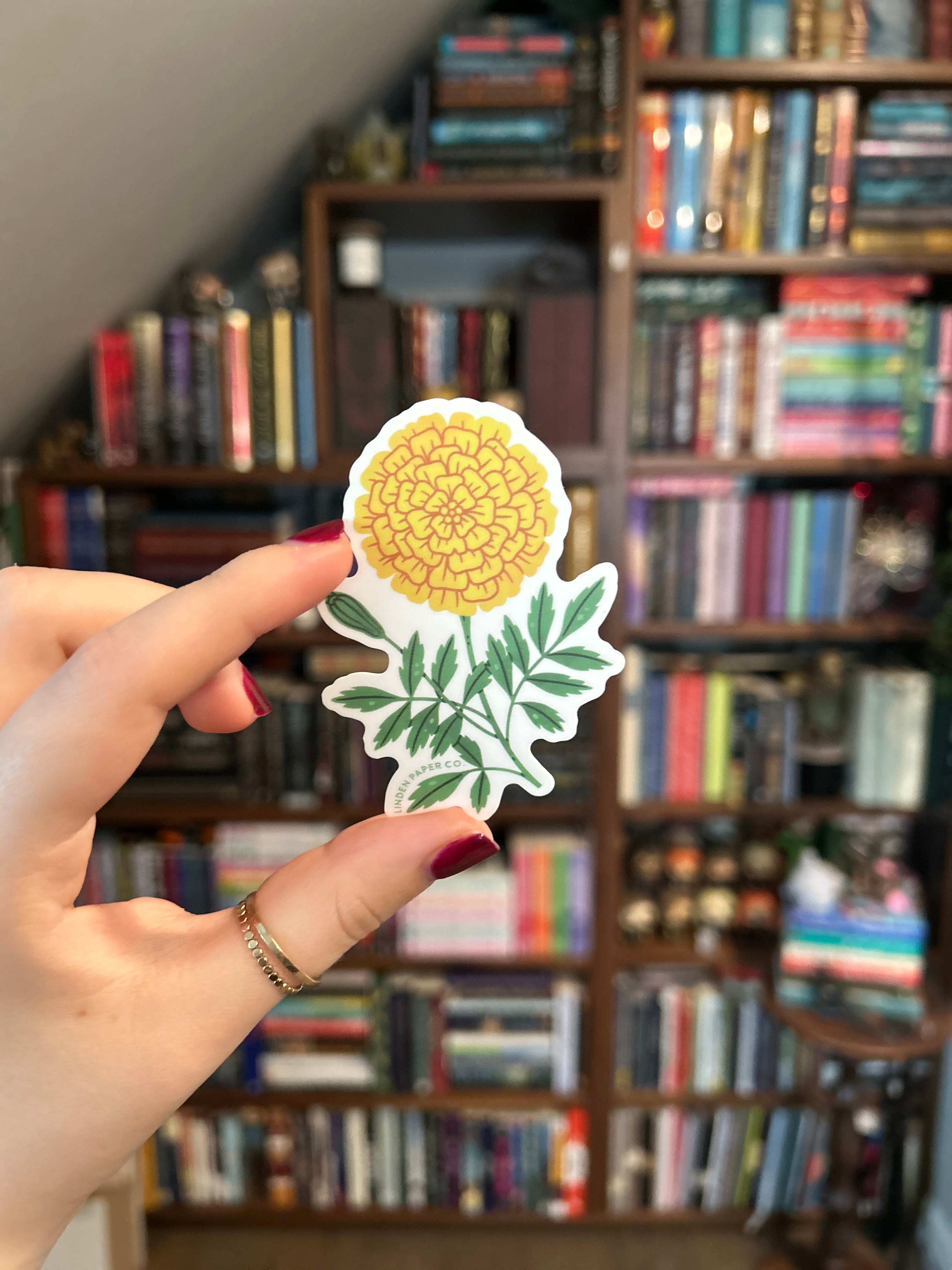 A hand with dark red nail polish holds a die-cut sticker featuring a yellow marigold flower and green leaves, with a blurred bookshelf in the background.