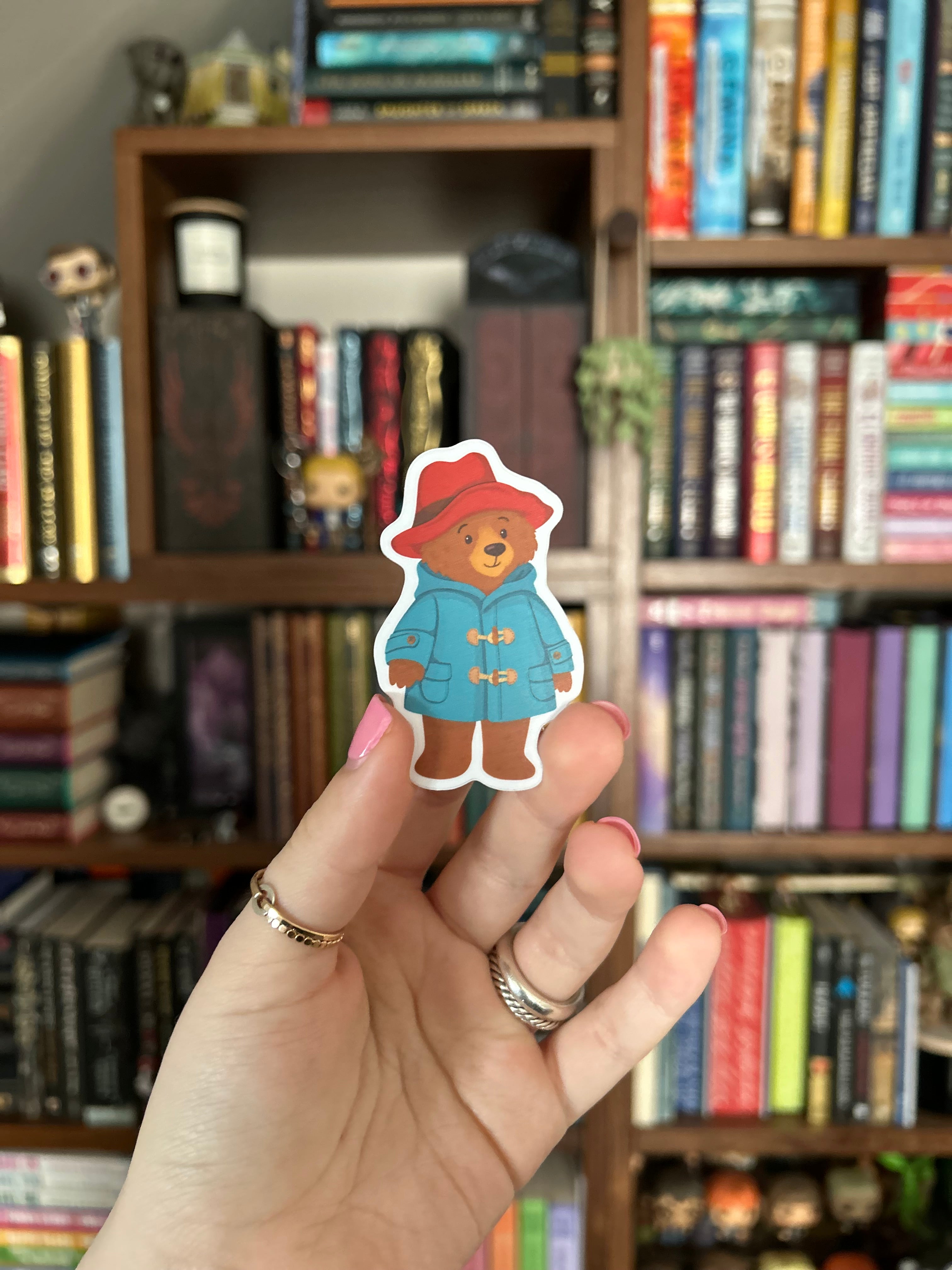 A hand holding a die-cut sticker of Marmalade Bear, with a blurred background of bookshelves filled with books.