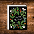 A Christmas greeting card with a black background and a colorful wreath design featuring green holly leaves, red berries, white flowers, pine branches, and the words 'Merry Christmas' in white script. The card is shown with a white envelope on a rustic brown wooden surface.