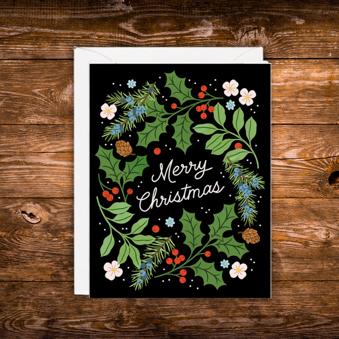 A Christmas greeting card with a black background and a colorful wreath design featuring green holly leaves, red berries, white flowers, pine branches, and the words 'Merry Christmas' in white script. The card is shown with a white envelope on a rustic brown wooden surface.
