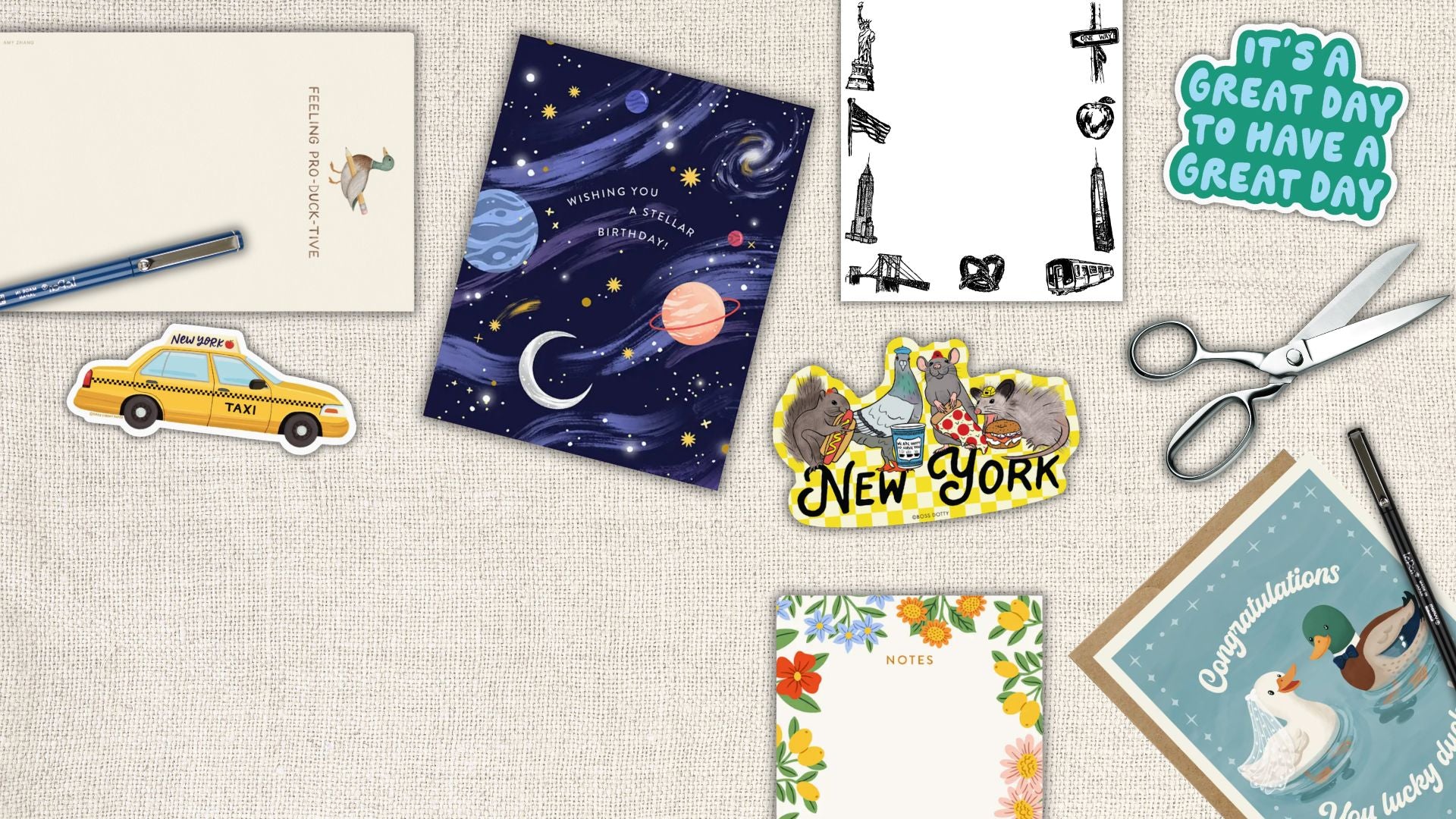 A flat-lay arrangement of stationery and craft items on a beige fabric background, including a New York-themed sticker sheet, a galaxy notepad, a yellow taxi sticker, scissors, a pen, and other paper goods.