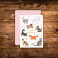 Mini birthday greeting card featuring playful illustrated cats surrounding the words "Happy Birthday" on a white background with a pink envelope.