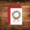 A mini Christmas greeting card with a white front featuring a green wreath decorated with red berries and a red bow, with the text 'merry christmas' below. A dark red envelope is placed behind the card on a rustic brown wooden surface.