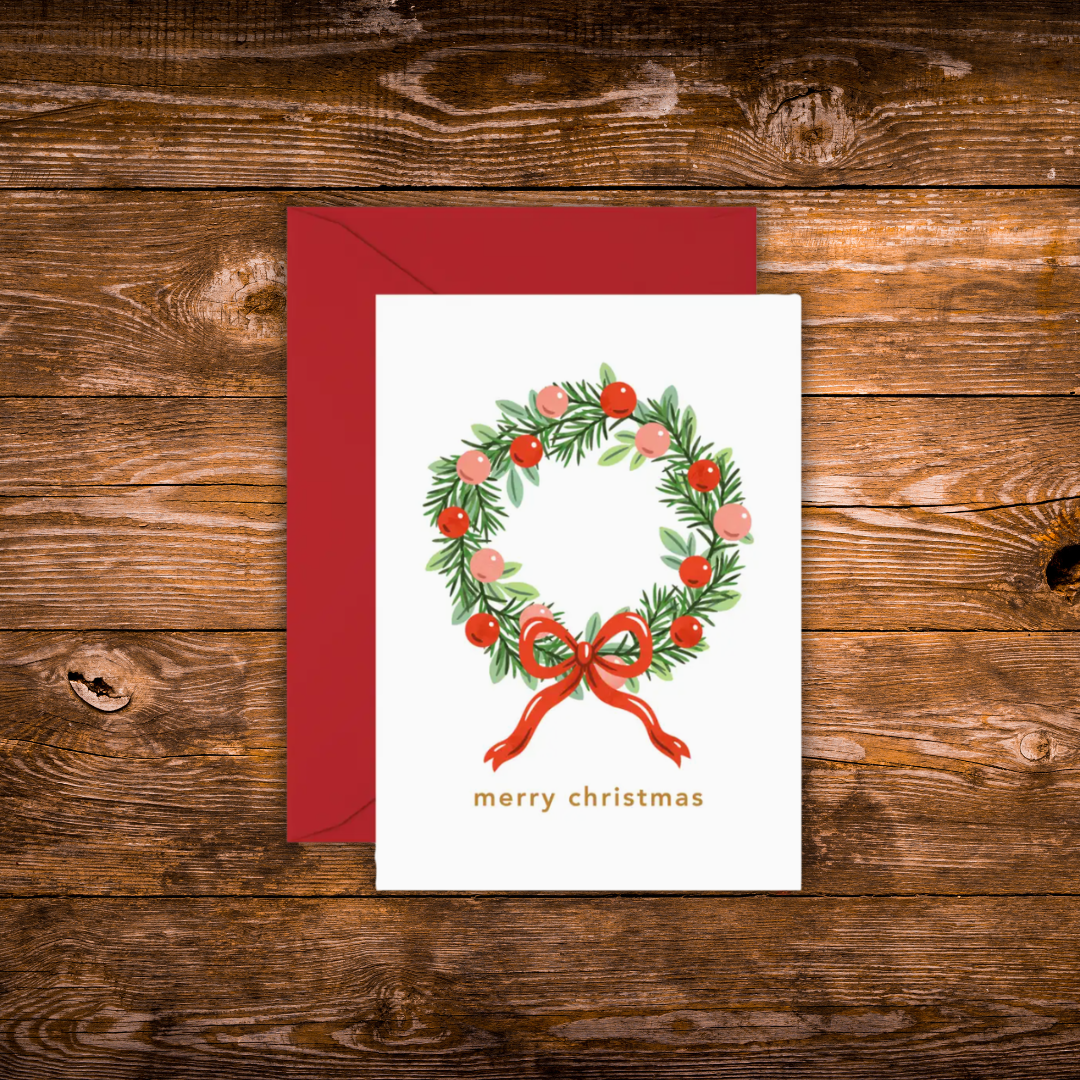 A mini Christmas greeting card with a white front featuring a green wreath decorated with red berries and a red bow, with the text 'merry christmas' below. A dark red envelope is placed behind the card on a rustic brown wooden surface.