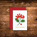 A mini white Christmas greeting card with a red envelope, featuring a holly branch with red berries and the text 'season's greetings' in gold, displayed on a rustic wooden background.