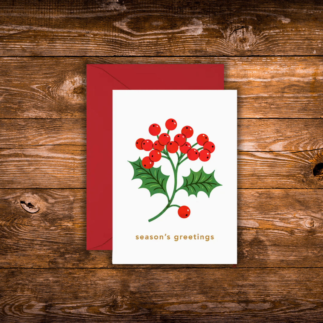 A mini white Christmas greeting card with a red envelope, featuring a holly branch with red berries and the text 'season's greetings' in gold, displayed on a rustic wooden background.