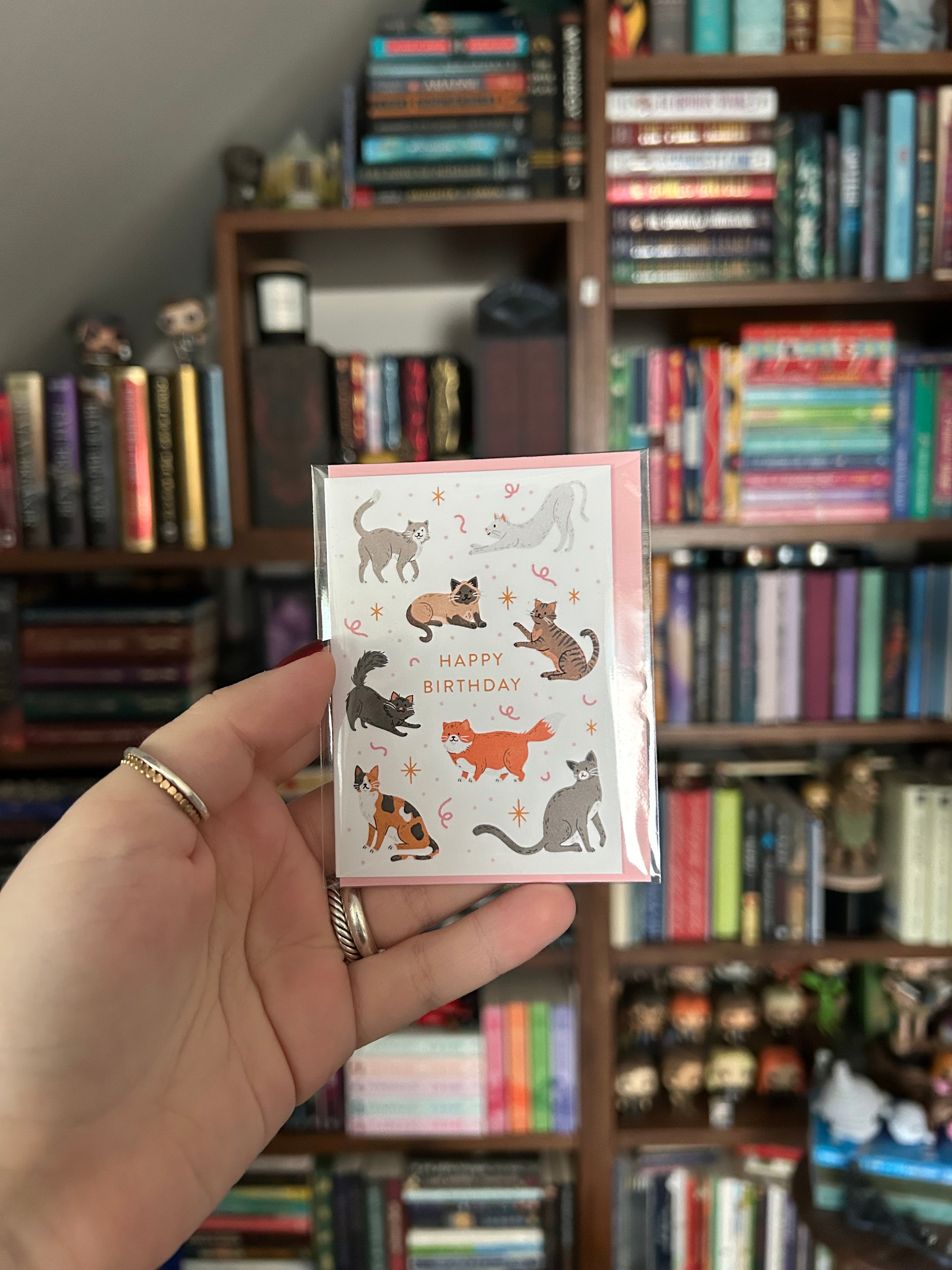 Mini birthday greeting card featuring playful illustrated cats surrounding the words "Happy Birthday" on a white background with a pink envelope.