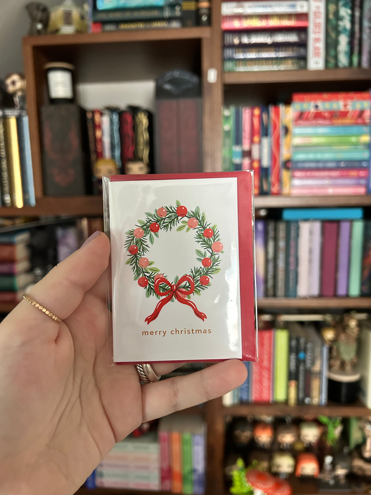 A hand holds a small Christmas greeting card featuring a green wreath with red berries and a red bow on a white background, with a blurred bookshelf behind it.