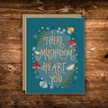 Whimsical woodland greeting card featuring mushrooms, wildflowers, moths, and stars with the hand-lettered message "There Is So Mushroom in My Heart for You" on a teal background.