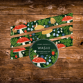 Linden Paper Co. Washi tape in a forest theme, featuring red and white mushrooms, blue and white flowers, and green foliage, shown with a round tin and several strips laid out on a rustic wooden background.