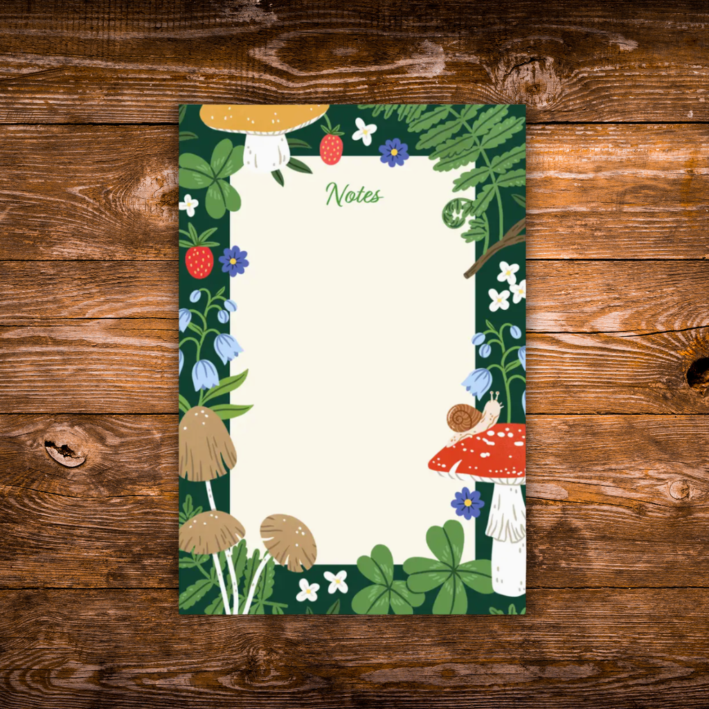 Woodland-themed notepad featuring mushrooms, ferns, strawberries, clovers, wildflowers, and a small snail surrounding a large blank writing area on a dark green background.