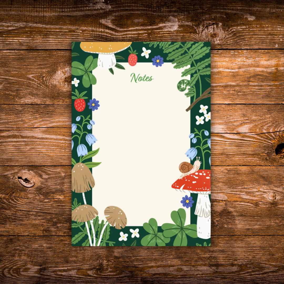 Woodland-themed notepad featuring mushrooms, ferns, strawberries, clovers, wildflowers, and a small snail surrounding a large blank writing area on a dark green background.