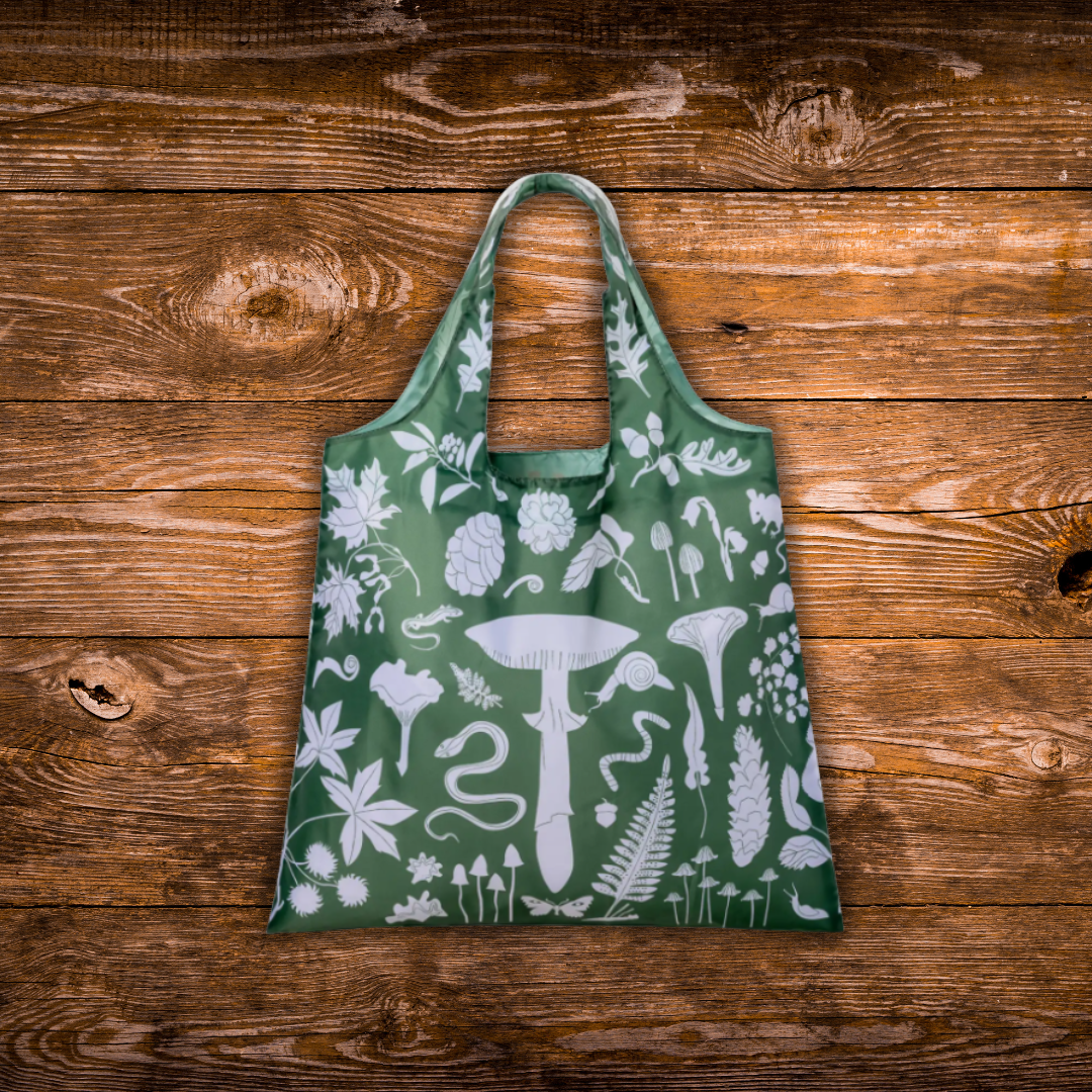 A green reusable tote bag with a white botanical and mushroom print, laid flat on a rustic brown wooden plank background.