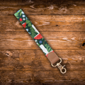 A fabric wristlet strap with a colorful woodsy and mushroom pattern, featuring a tan leather loop and a gold metal swivel clasp, displayed diagonally on a rustic brown wooden plank background.