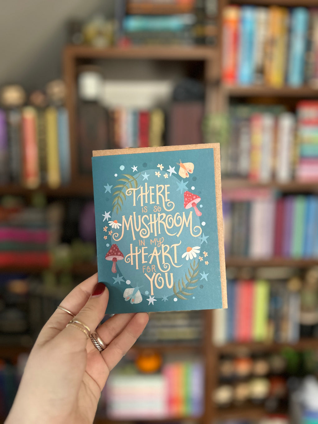 Whimsical woodland greeting card featuring mushrooms, wildflowers, moths, and stars with the hand-lettered message "There Is So Mushroom in My Heart for You" on a teal background.