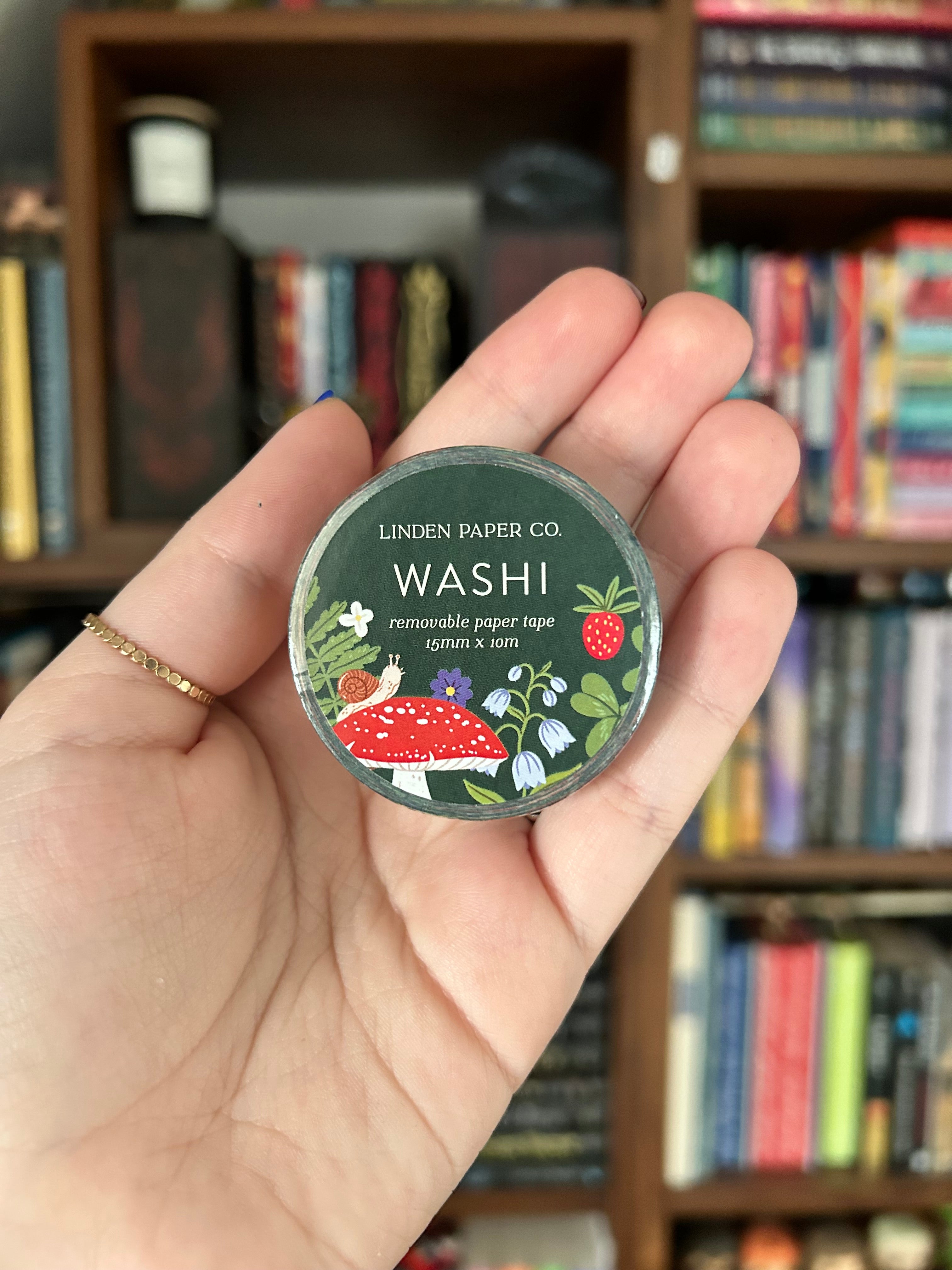 A hand holding a washi tape from Linen Paper Co. featuring a forest-themed label with red strawberries, green leaves, and a red-and-white spotted fox, set against a blurred background of bookshelves.