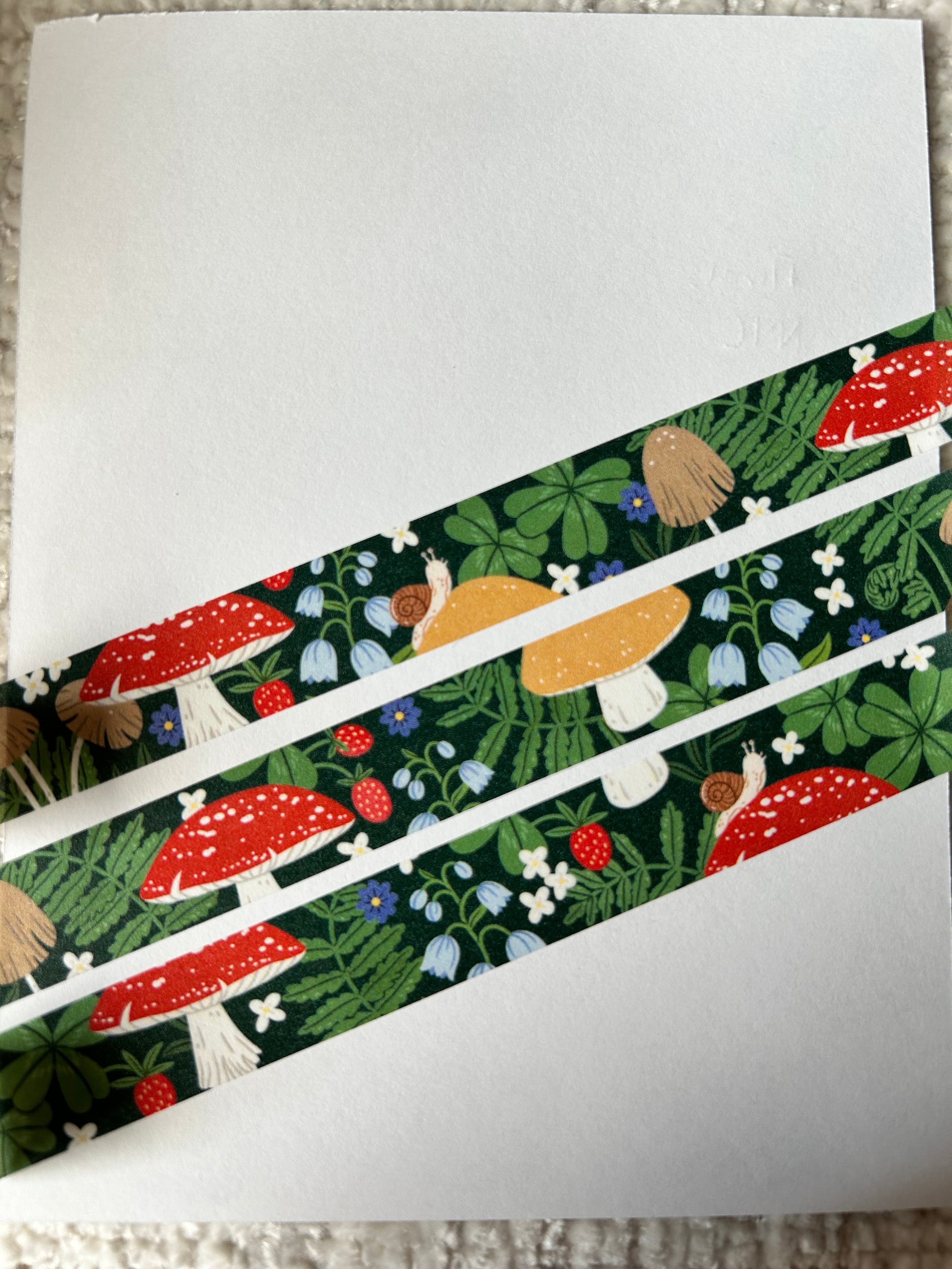 A close-up of a white card or sheet with three diagonal strips of patterned washi tape featuring a forest design with red and yellow mushrooms, green leaves, and small blue and white flowers.
