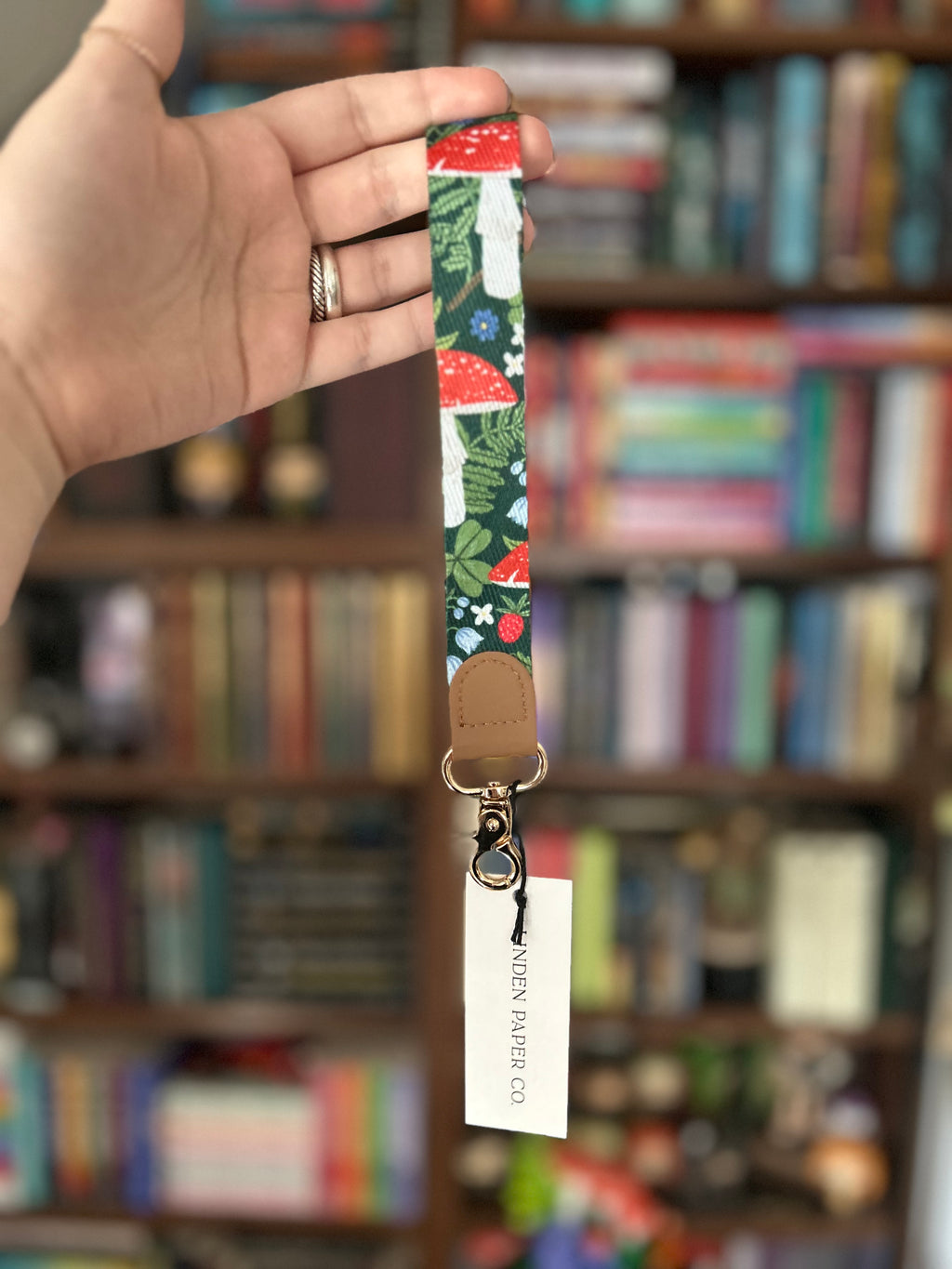 A hand holds a fabric lanyard with a colorful mushroom and floral print, a tan leather loop, and a white hangtag that reads 'LINDEN PAPER CO.' in front of a blurred bookshelf background.