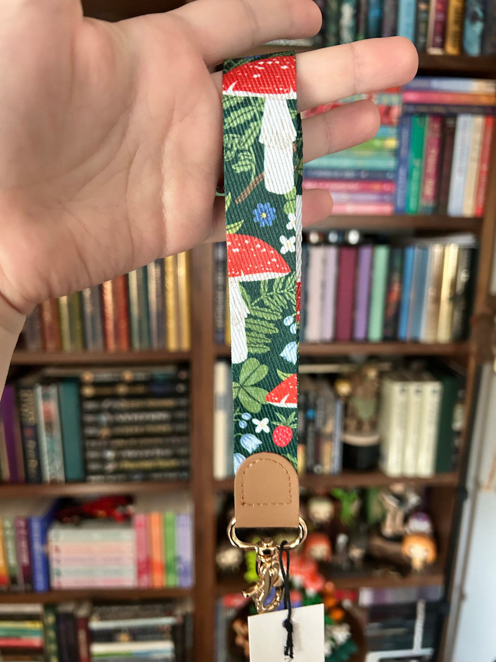 A close-up of a hand holding a fabric wrist strap with a colorful mushroom and floral pattern, a tan leather tab, and a gold-tone metal ring, with a blurred bookshelf in the background.