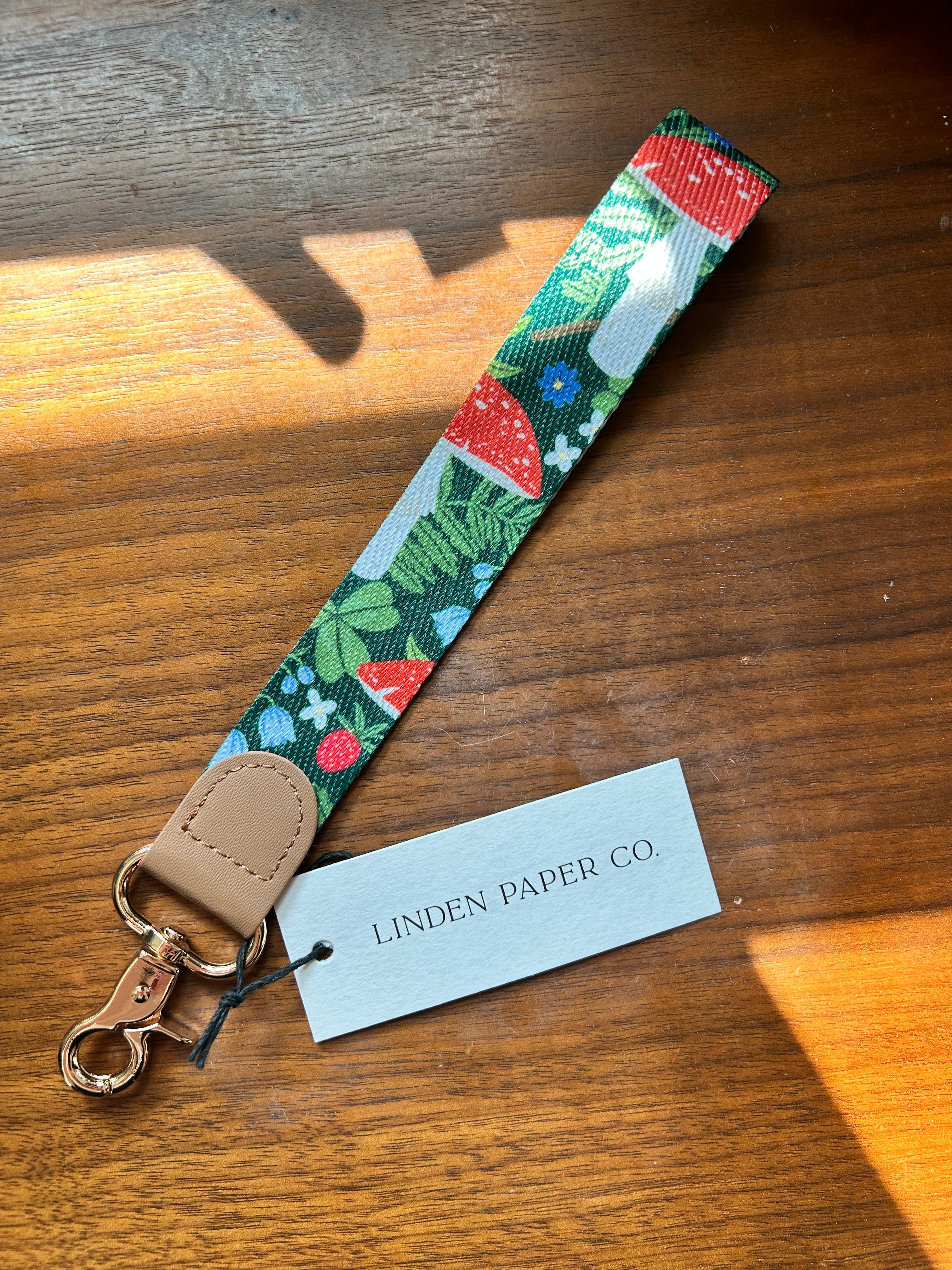 A fabric wristlet strap with a colorful mushroom and leaf print, featuring a tan leather loop and a gold-tone clasp, shown on a wooden table next to a Linden Paper Co. paper tag.
